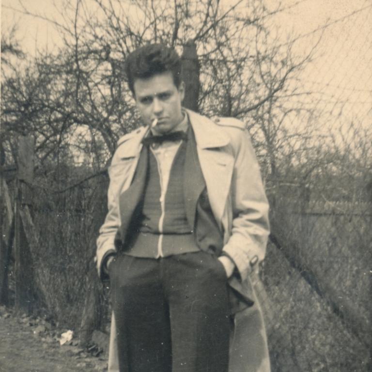 Tommy steht vor dem Garten an der Meyerbeerstraße. Sie trägt einen hellen Trenchcoat, Fliege und eine Haartolle im Stil von Elvis Presley. 